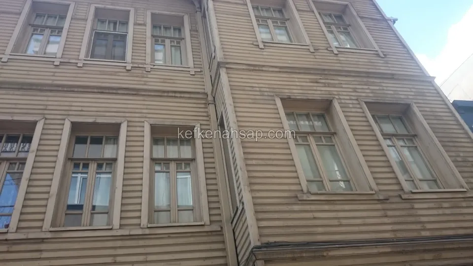 Alttan görünen, bej boyalı yatay ahşap kaplamalı üç katlı bir binanın düzenli sıralı pencereli cephesi