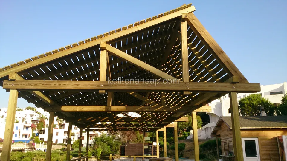 Beyaz evlerin önünde, kalın dikmeler üzerinde yükselen beşik çatılı, çıtalı ahşap pergola
