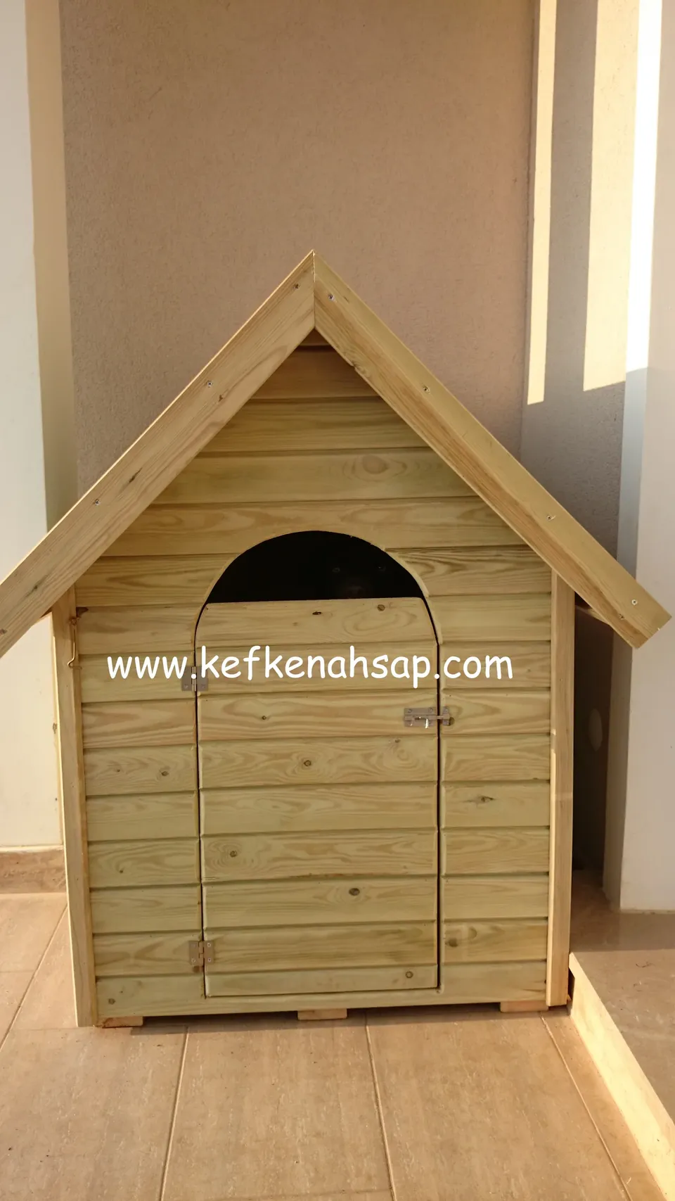Kemer biçimli girişi olan, yatay tahtalı ahşap köpek kulübesi karo zeminde duruyor; arkada düz bir duvar var