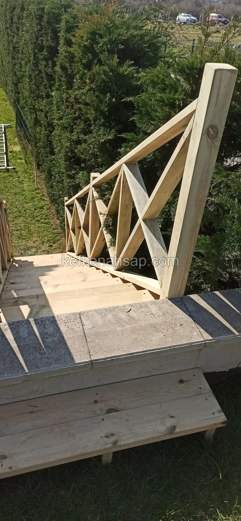 Beton basamağın üstünde ahşap platform ve çaprazlı korkuluk; arkada sık selvi çiti