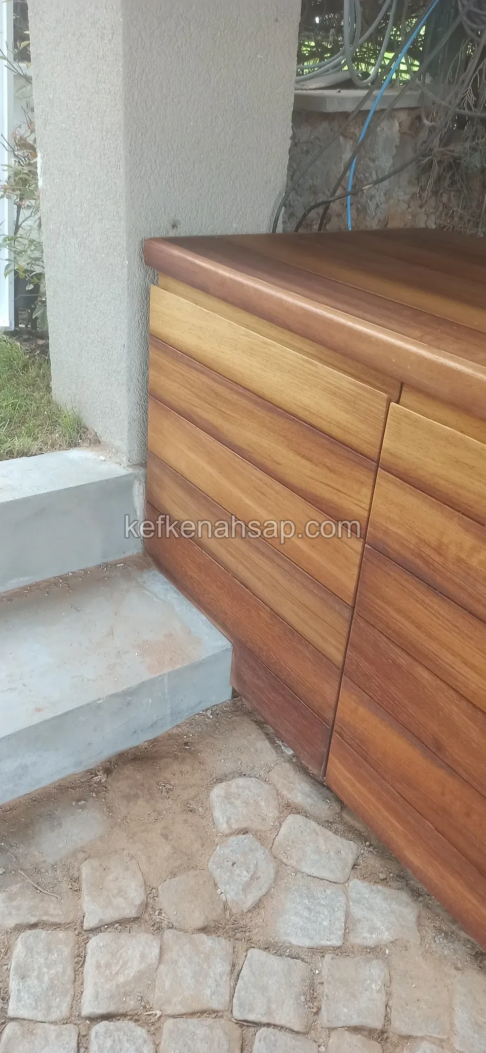Ein zweitüriger Holzschrank mit waagerechten Paneelen auf Kopfsteinpflaster neben einer Betonstufe