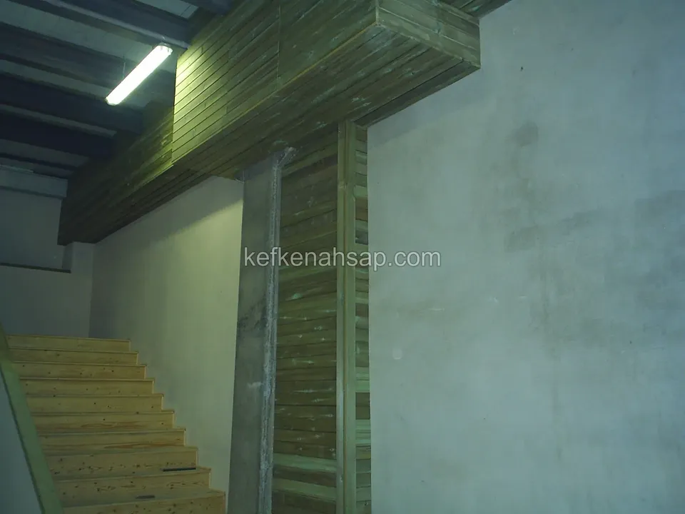 Beton duvarlar arasında açık renkli ahşap basamaklı iç merdiven; yanında tahta kaplanmış kolon ve kiriş