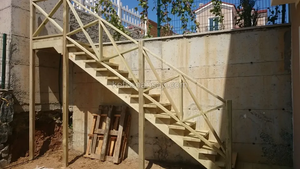 Beton istinat duvarı boyunca uzanan çaprazlı korkuluklu ahşap merdiven; zeminde paletler, arkada evler