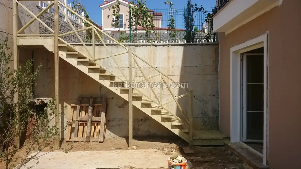 Beton duvar boyunca çaprazlı korkuluklu ahşap merdiven; sağda evin kapısı, zeminde toprak ve palet