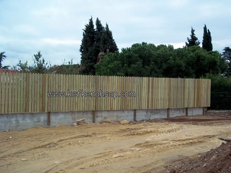 Alçak beton duvarın üstünde uzanan, dikey tahtalı uzun ahşap çit ve arkasında ağaçlar