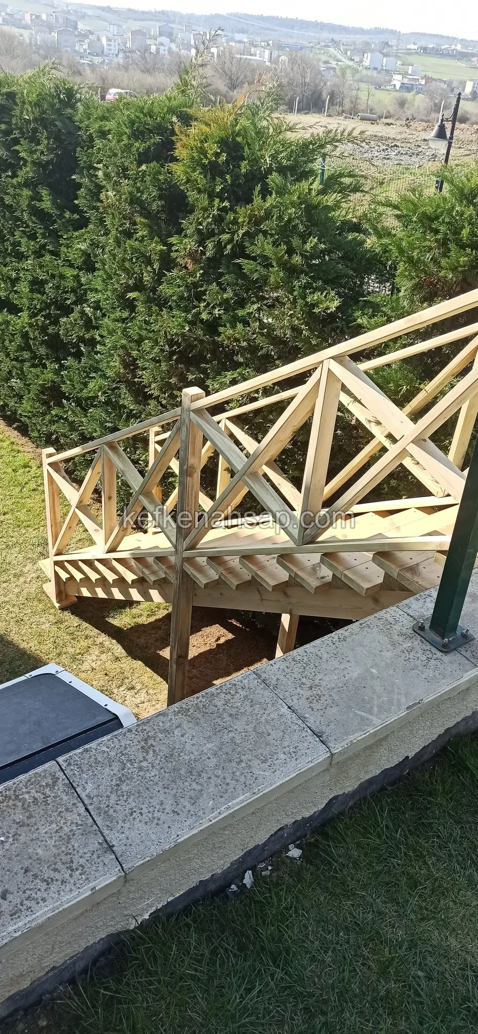 Holzpodest auf Stützen mit Kreuzstreben-Geländer an einer Betonkante, dahinter Bäume und Fernblick