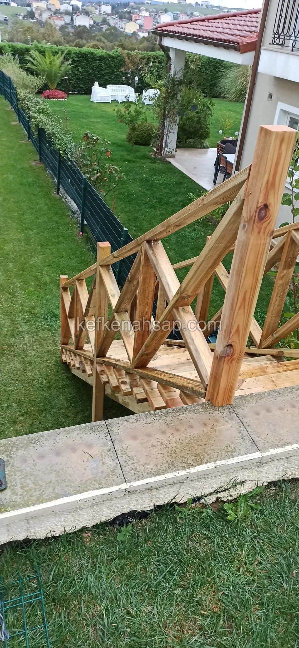 Holztreppe mit Kreuzstreben-Geländer, die von einer Betonkante in einen Rasengarten hinabführt