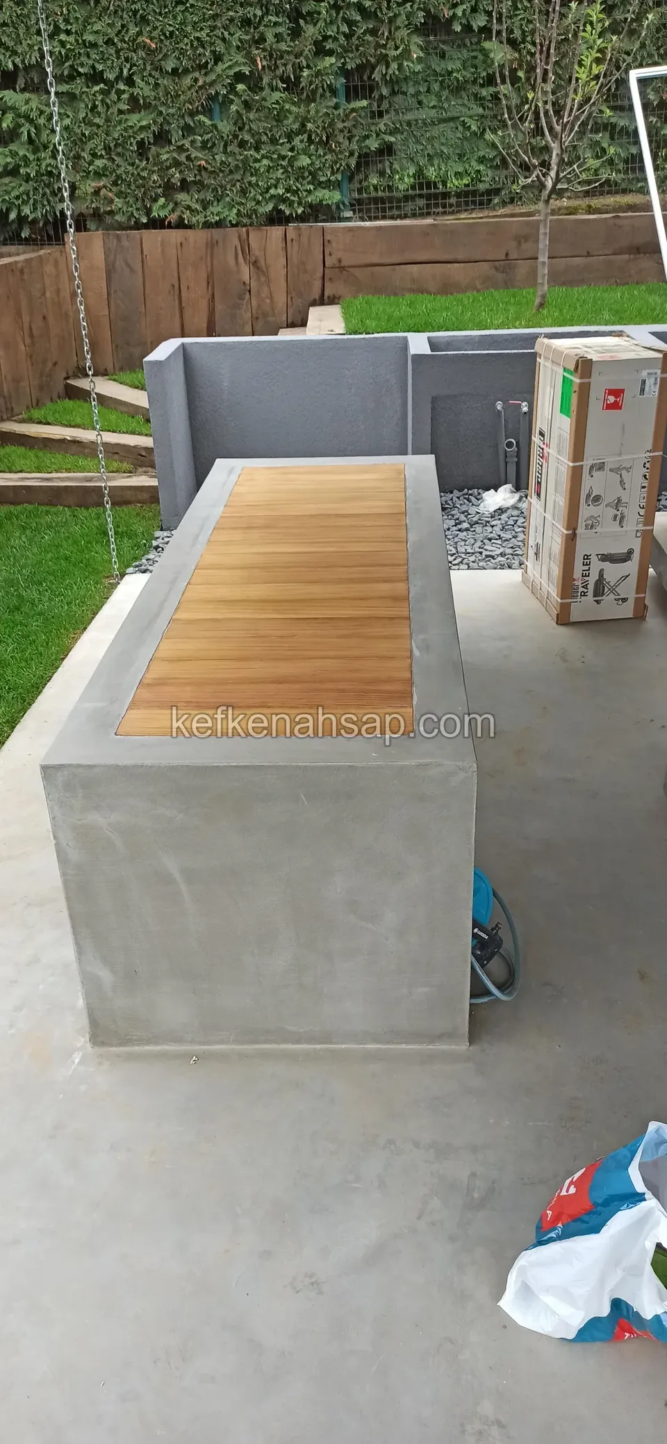 Gri beton tezgâhın ortasında ahşap çıtalı yüzey; arkada ambalajlı bir koli, çim bahçe ve gri duvar