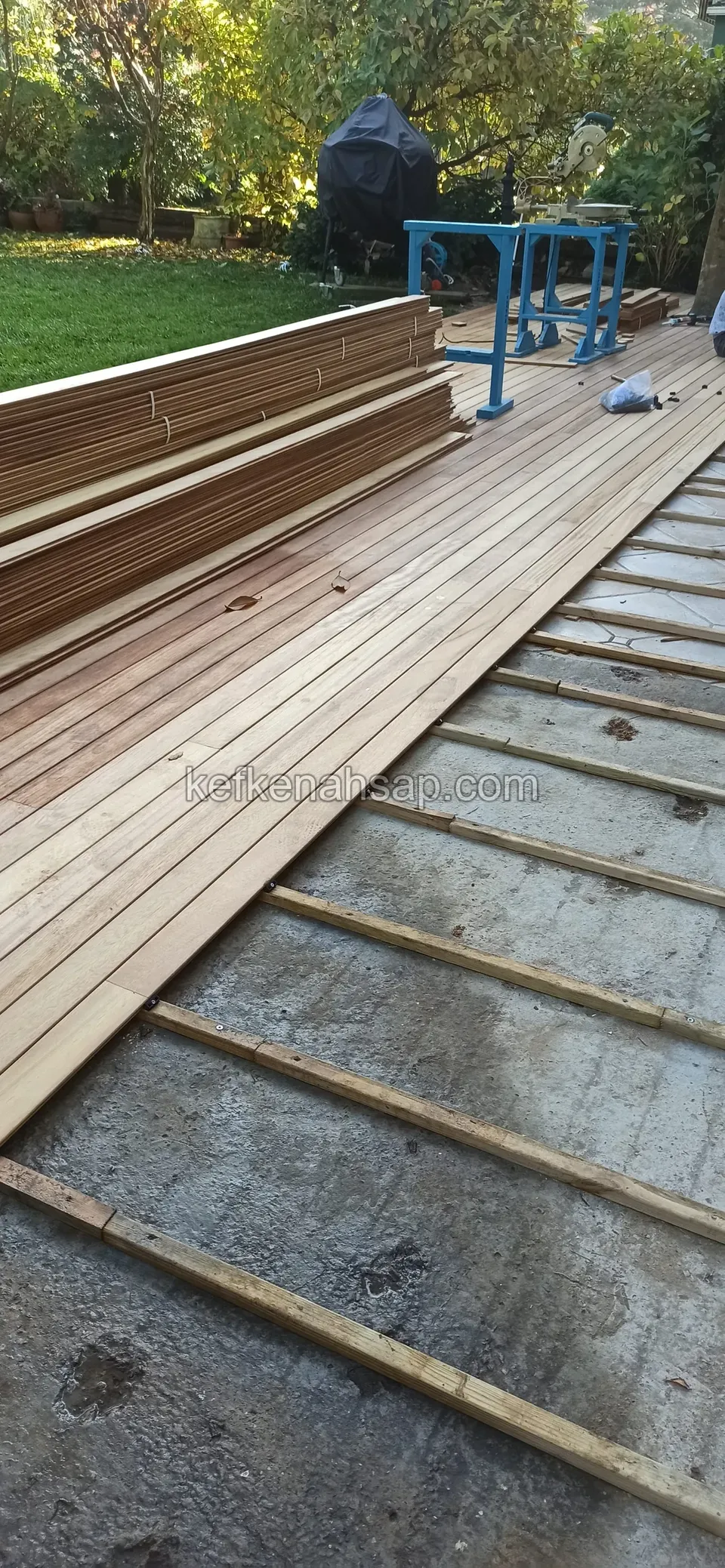 Latten auf einem Betonboden, ein Teil des Decks schon verlegt, am Rand ein Holzstapel und ein blauer Bock