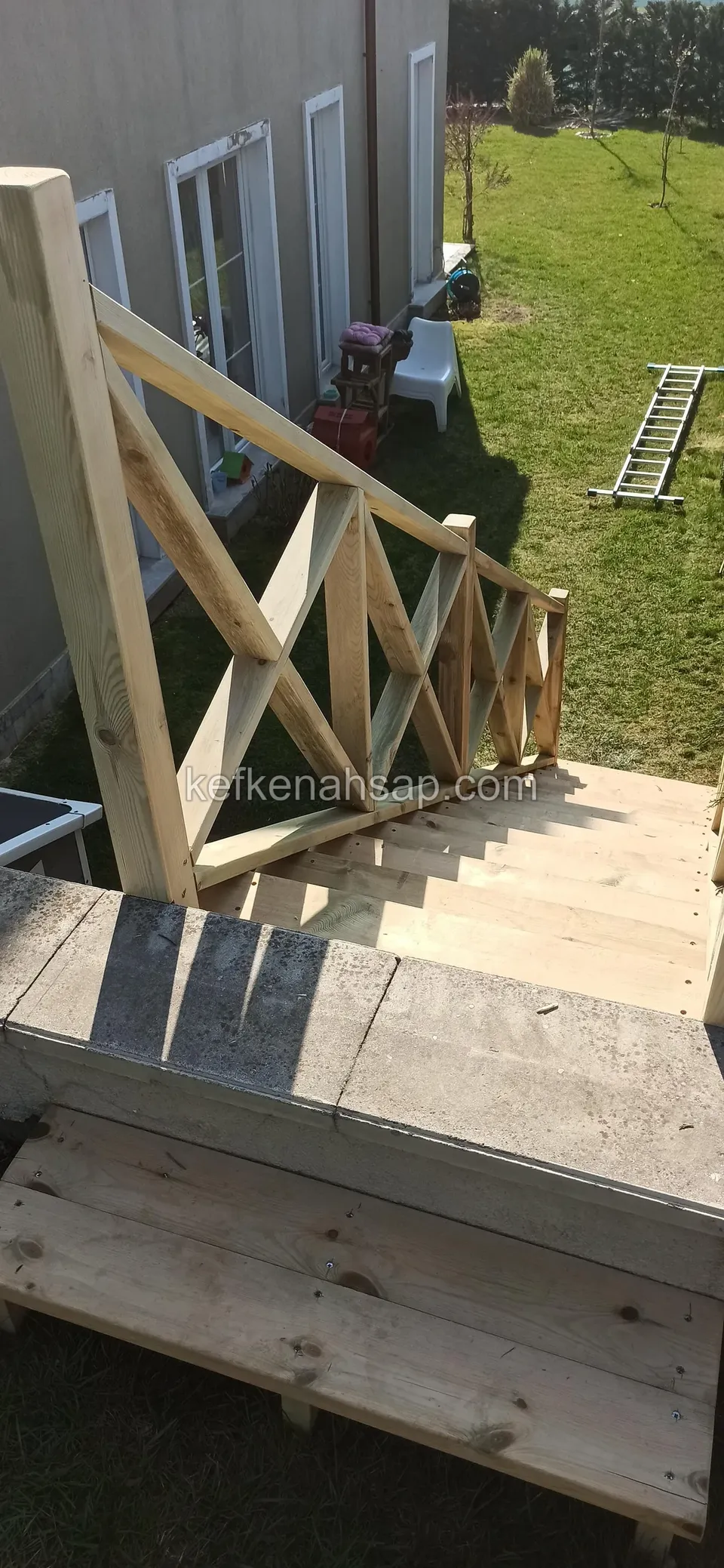 Betonkante und Podest mit Holzstufe, Kreuzstreben-Geländer, dahinter ein Haus mit Fenstern und Rasen