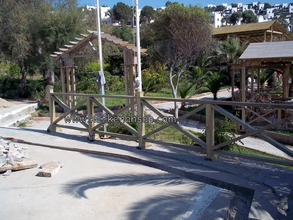 Beton yolun kenarında uzanan çapraz çıtalı açık tonlu ahşap korkuluk, arkada pergola ve ahşap çatılı bir kameriye