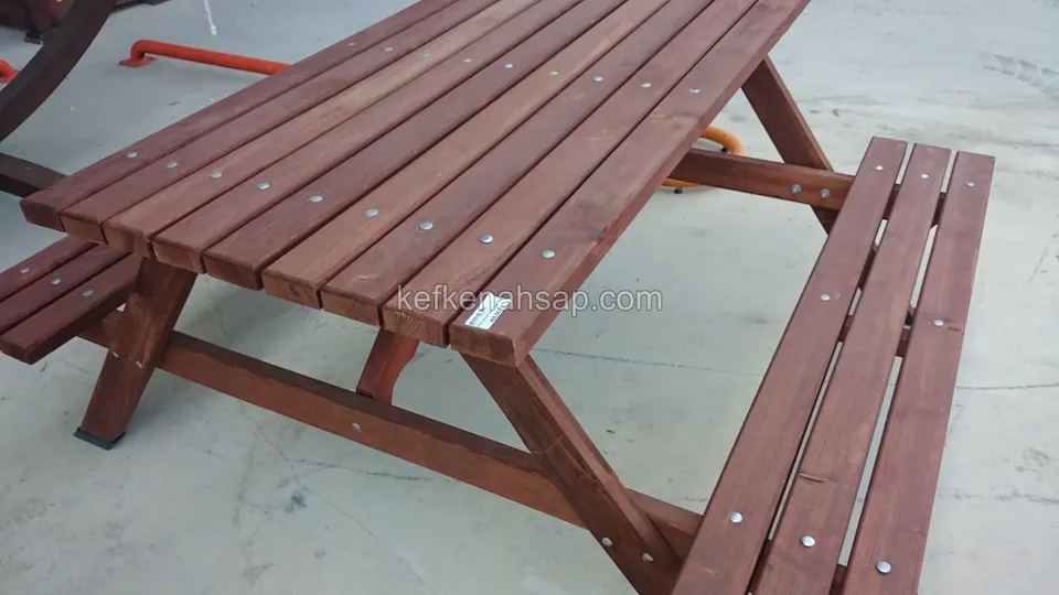 Beton zeminde duran, kırmızımsı kahverengi boyalı, çaprazlanan ayaklı ve bağlı bankları olan ahşap piknik masası