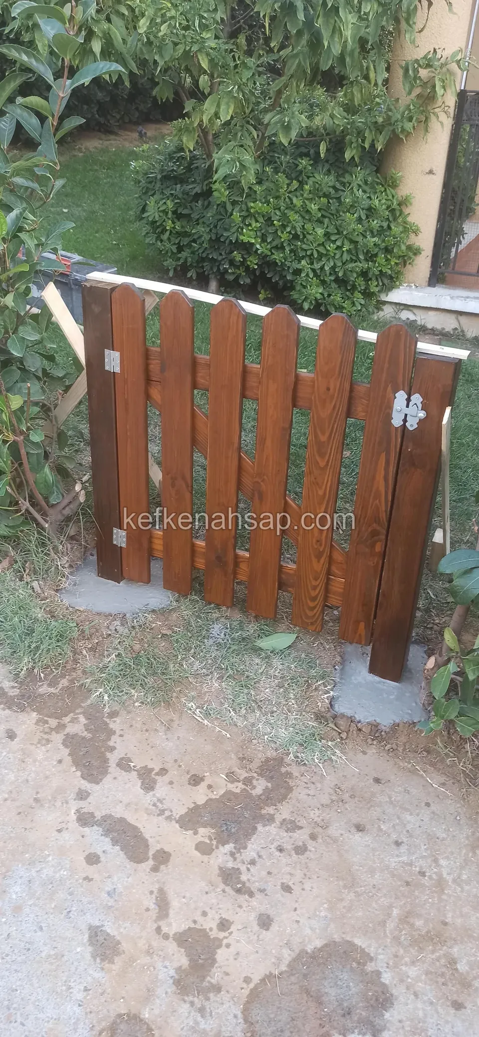 Beton zemine metal ayaklarla sabitlenmiş, kahverengi cilalı çıtalı kapı, arkada yeşillik