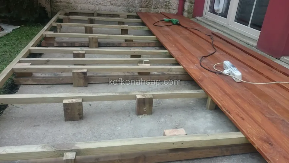 Beton zeminde ahşap takozlara oturan kiriş iskeleti, sağ tarafta kızıl kahve deck tahtaları döşenmiş, yerde kablo