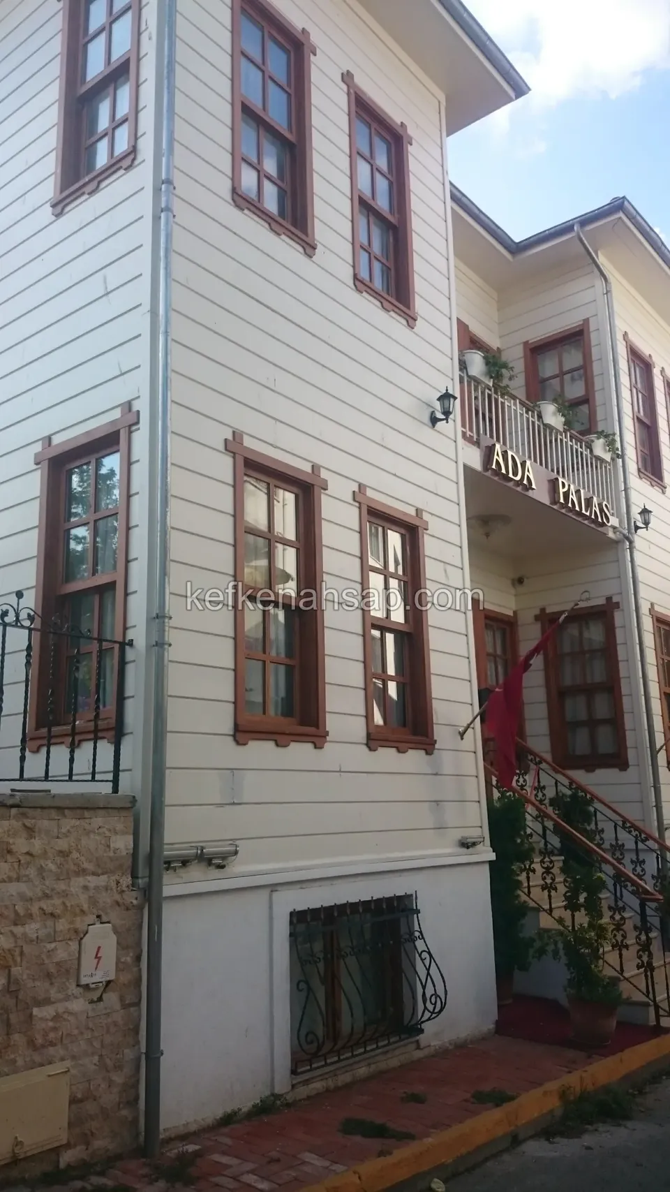 Beyaz boyalı yatay kaplamalı çok katlı ahşap bina; kahverengi pencere doğramaları, üstte balkon ve altta taş duvar