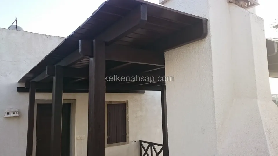 Beyaz bir binanın girişinde, koyu vernikli kalın direklerle taşınan düz çatılı ahşap sundurma