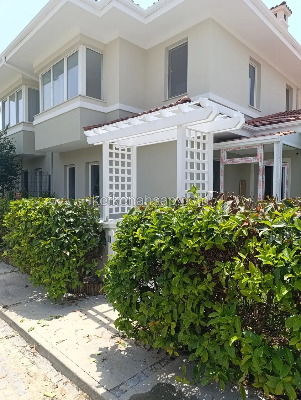 Villa girişinde beyaz boyalı ahşap pergola, kafes yan paneller ve iki yanda budanmış çalılar