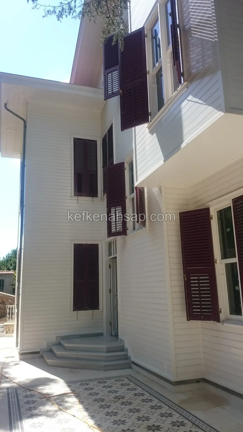 Beyaz boyalı yatay ahşap kaplamalı binada bordo panjurlar ve girişte taş basamaklar