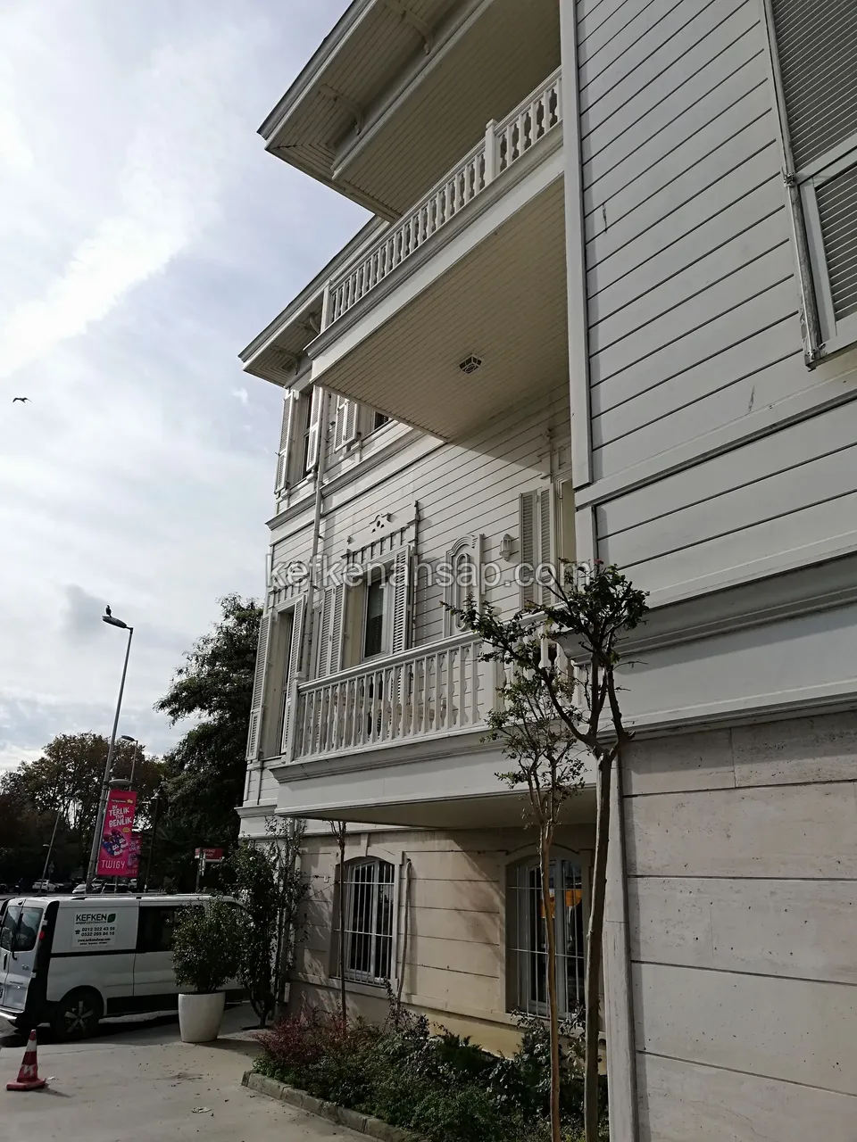 Sokak köşesinde, beyaz boyalı yatay ahşap kaplamalı çok katlı bina cephesi ve işlemeli balkon korkulukları