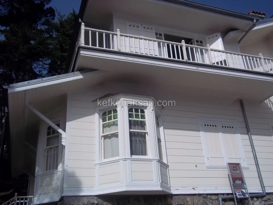 Beyaz boyalı ahşap kaplamalı yapıda balkon korkuluğu, altında çıkma cumbalı pencere ve yanında kepenkli bir pencere