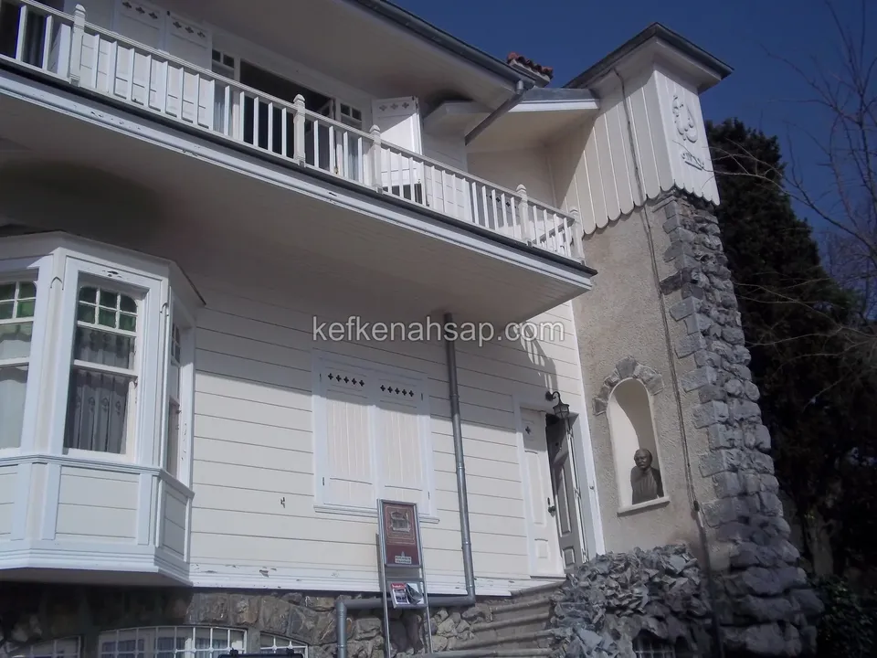 Beyaz boyalı ahşap kaplamalı yapının balkonu ve cumbalı penceresi, sağda taşla örülmüş bir kule ve önünde taş basamaklar