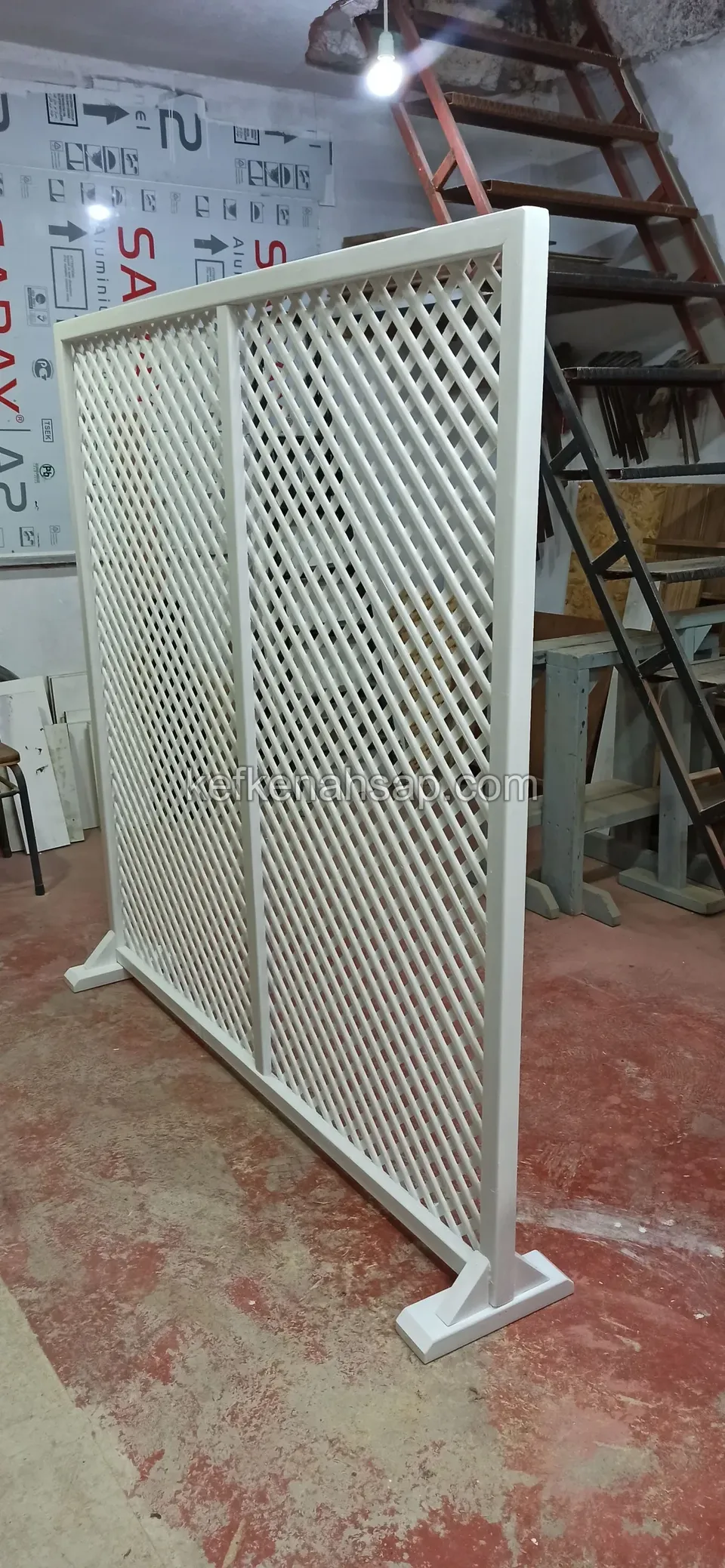 Atölyede ahşap merdivenin önünde, kırmızımsı betonda duran beyaz boyalı çaprazlı kafes panel; sol ayağı yere basıyor
