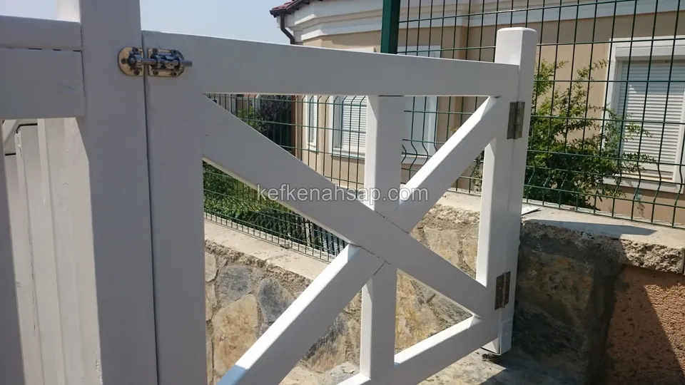 Beyaz boyalı çapraz kirişli ahşap kapı kanadı, siyah mandalı ve menteşesi yakından; arkada tel çit ve taş duvar
