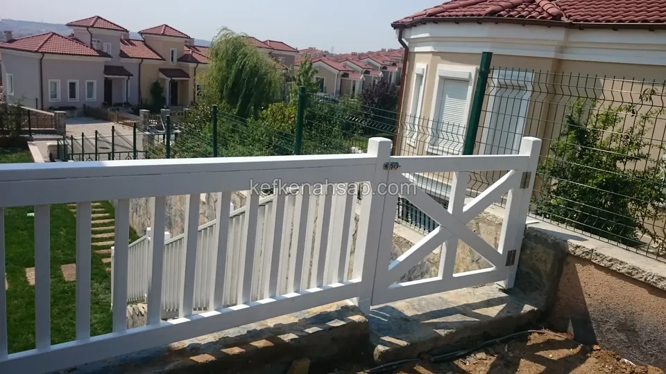 Taş duvarın üstünde beyaz boyalı dikey çıtalı korkuluk ve ortasında X çaprazlı kapı, arkada tel çit ve kiremit çatılı evler