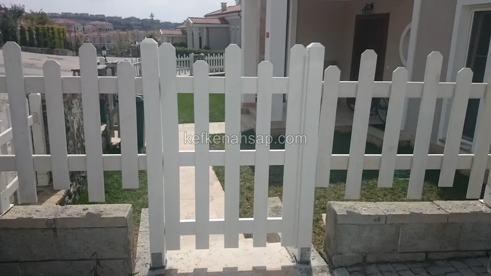 İki taş blok arasındaki giriş boşluğuna oturan beyaz boyalı çıtalı kapı ve iki yana uzanan beyaz çit, arkada evler