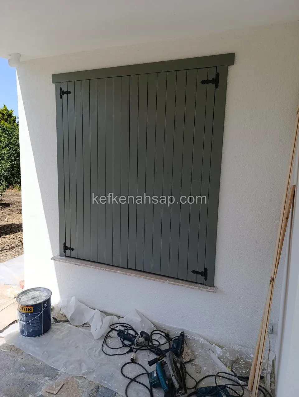 Beyaz cephede pencereyi kapatan adaçayı yeşili dikey çıtalı ahşap panjur, yerde boya kovası ve koruyucu örtü