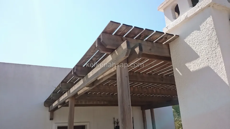 Beyaz sıvalı binanın girişinde, koyu tonlu kirişleri ve çıtalı üst örtüsüyle duvara bağlanan pergola
