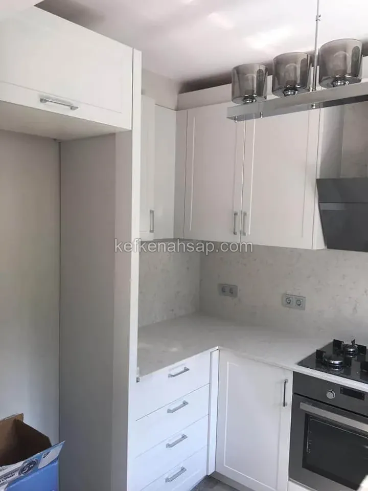 Beyaz çerçeveli kapaklı mutfak dolapları, mermer desenli tezgah ve ocak, fırın ile gri camlı sarkıt lambalar
