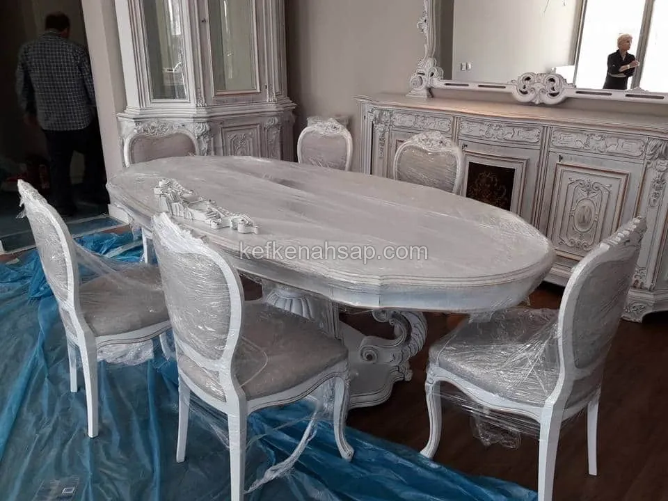 White oval dining table with carved details, upholstered chairs, display cabinet and buffet, covered in protective plastic