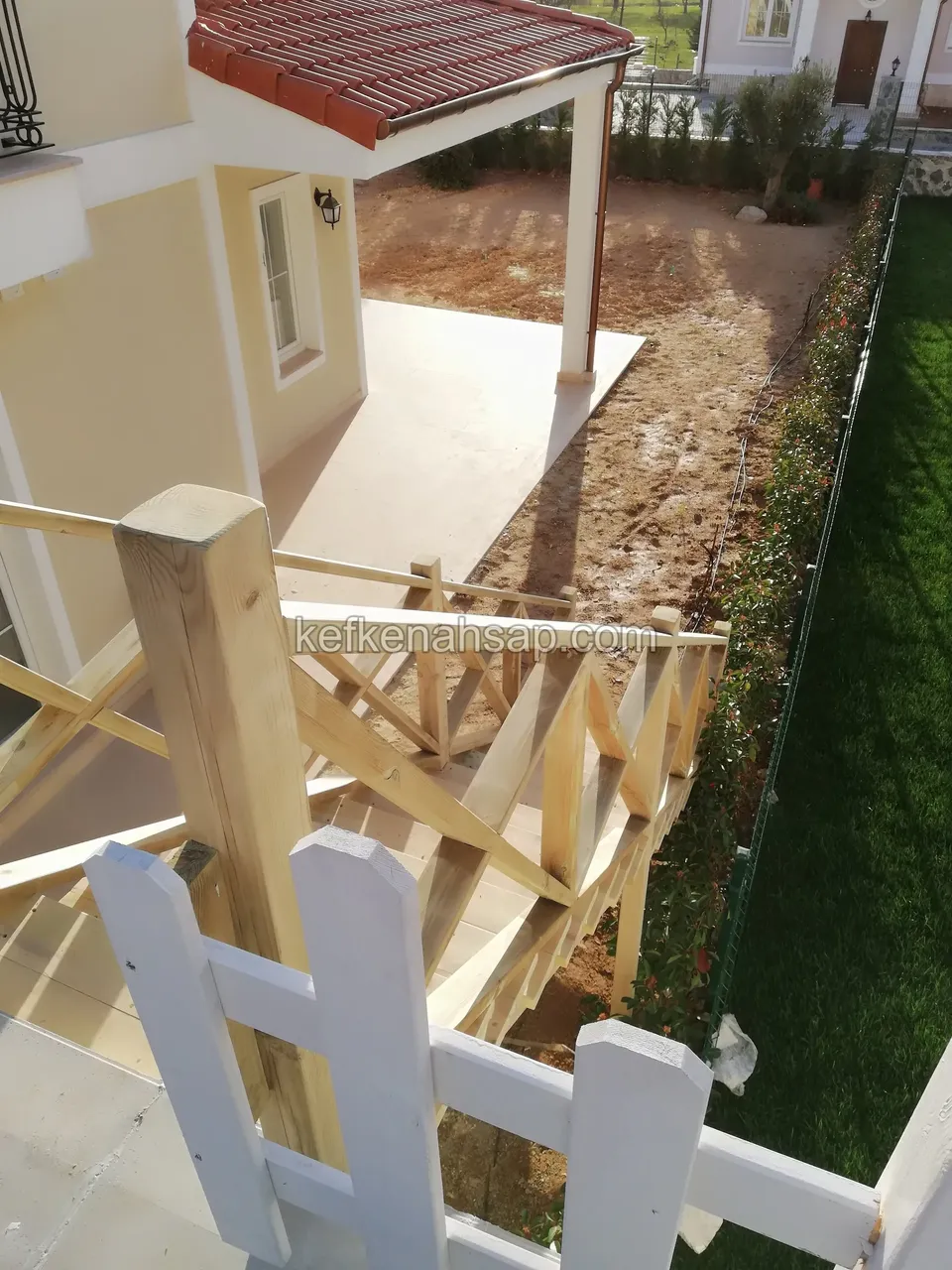 Kırmızı çatılı sarı evin yanında, yukarıdan görünen X çaprazlı ahşap korkuluk ve önde beyaz boyalı çıtalı çit, aşağıda çimlik