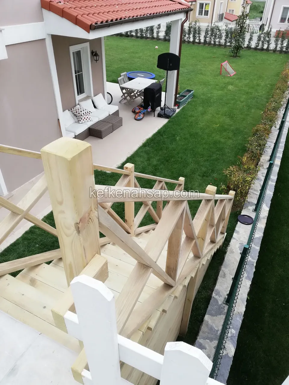 Holztreppe mit Kreuzstreben-Geländer neben einem weißen Zaunpfosten, dahinter Rasen, Terrasse und Sitzgruppe