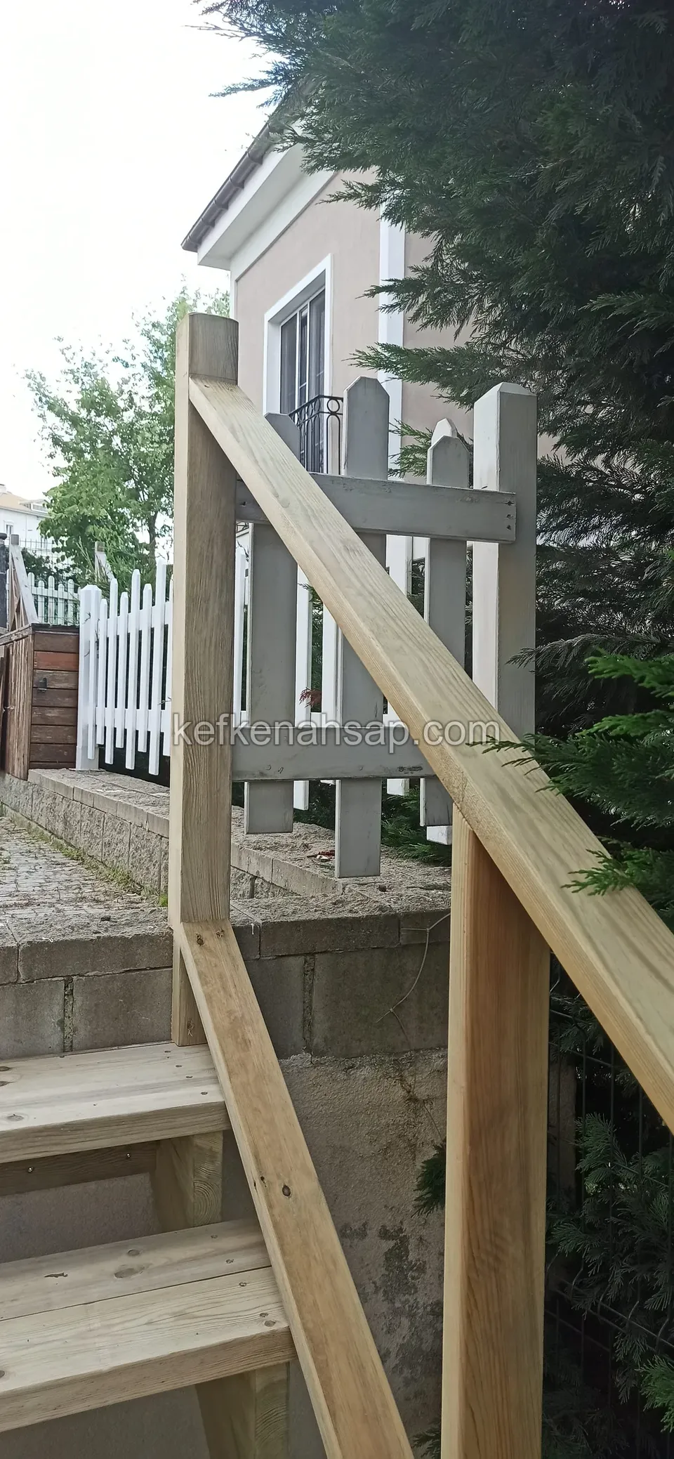 Helle Holztreppe mit Handlauf neben einem weißen Zaun, dahinter die Fassade eines rosa Hauses