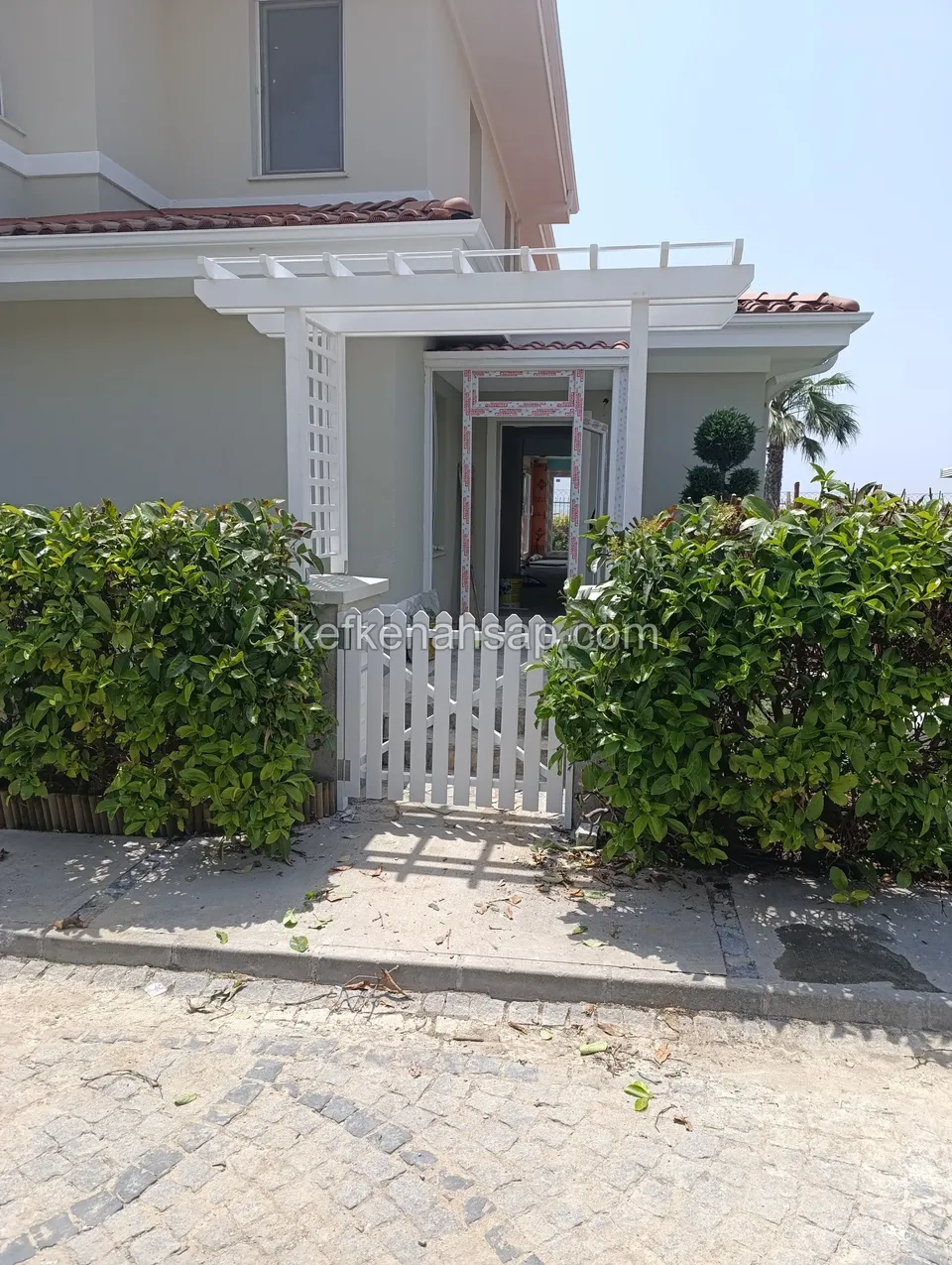 Çit bitkileri arasında beyaz çıtalı ahşap bahçe kapısı ve arkasında kafes yan panelli beyaz pergola ile villa girişi