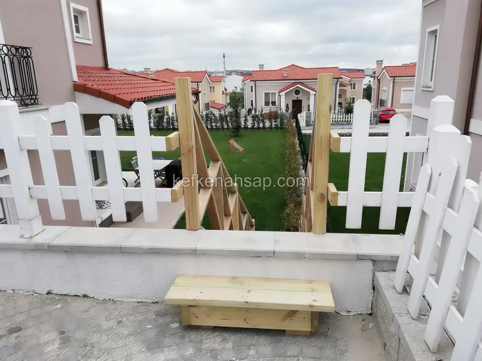 Çim bahçeyi çeviren beyaz boyalı alçak çit ve alçak duvarın üzerine kurulan, çapraz destekli ahşap kapı iskeleti