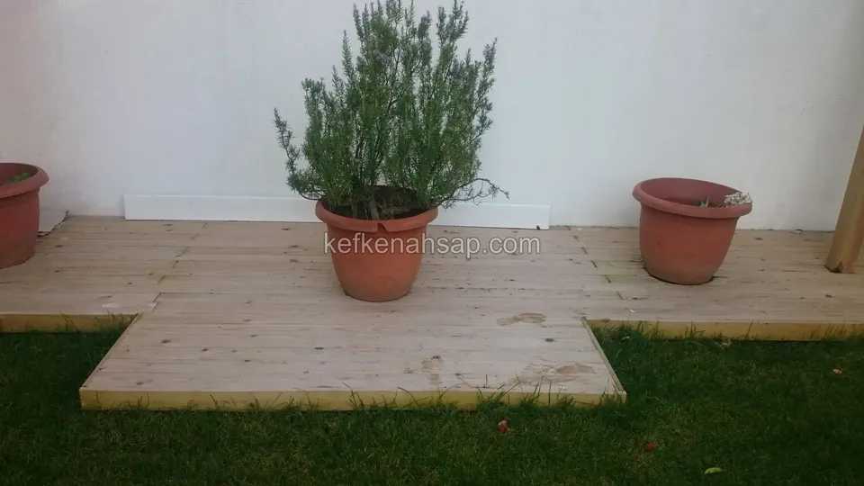 Beyaz duvarın dibinde çimle sınırlanan açık renkli deck platformu, üzerinde biberiye ve iki kırmızı toprak saksı