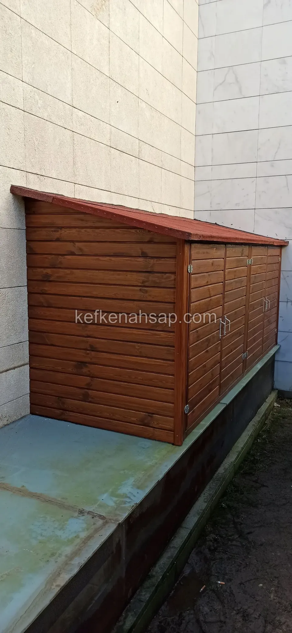 Niedriger Holzschrank mit waagerechten Brettern und rotem Pultdach auf einem Betonsockel an einer weißen Wand