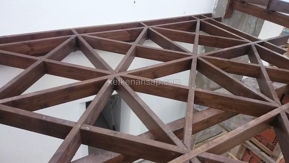 Beyaz duvarın önünde üçgen geometrik desen oluşturan vernikli ahşap pergola kafesi, köşede tuğla kolon