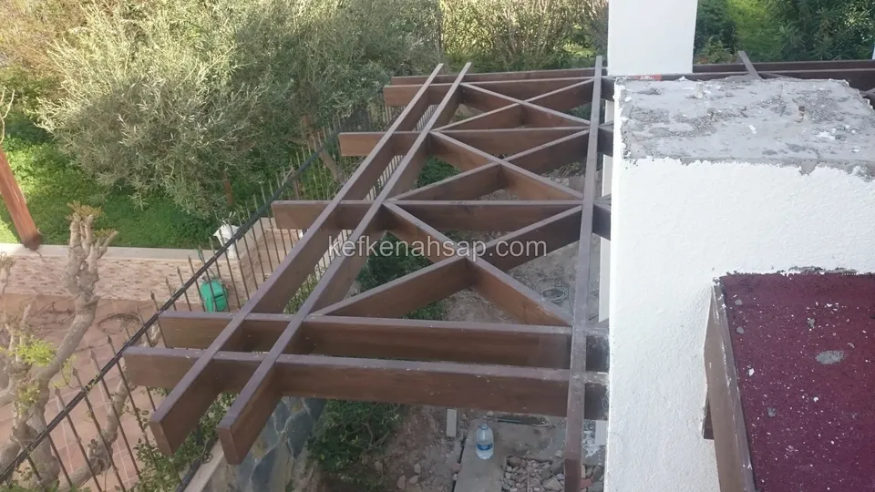 Beyaz duvarın yanında üçgen desenli vernikli pergola kafesinin kenarı, altında yeşil bahçe ve demir korkuluk