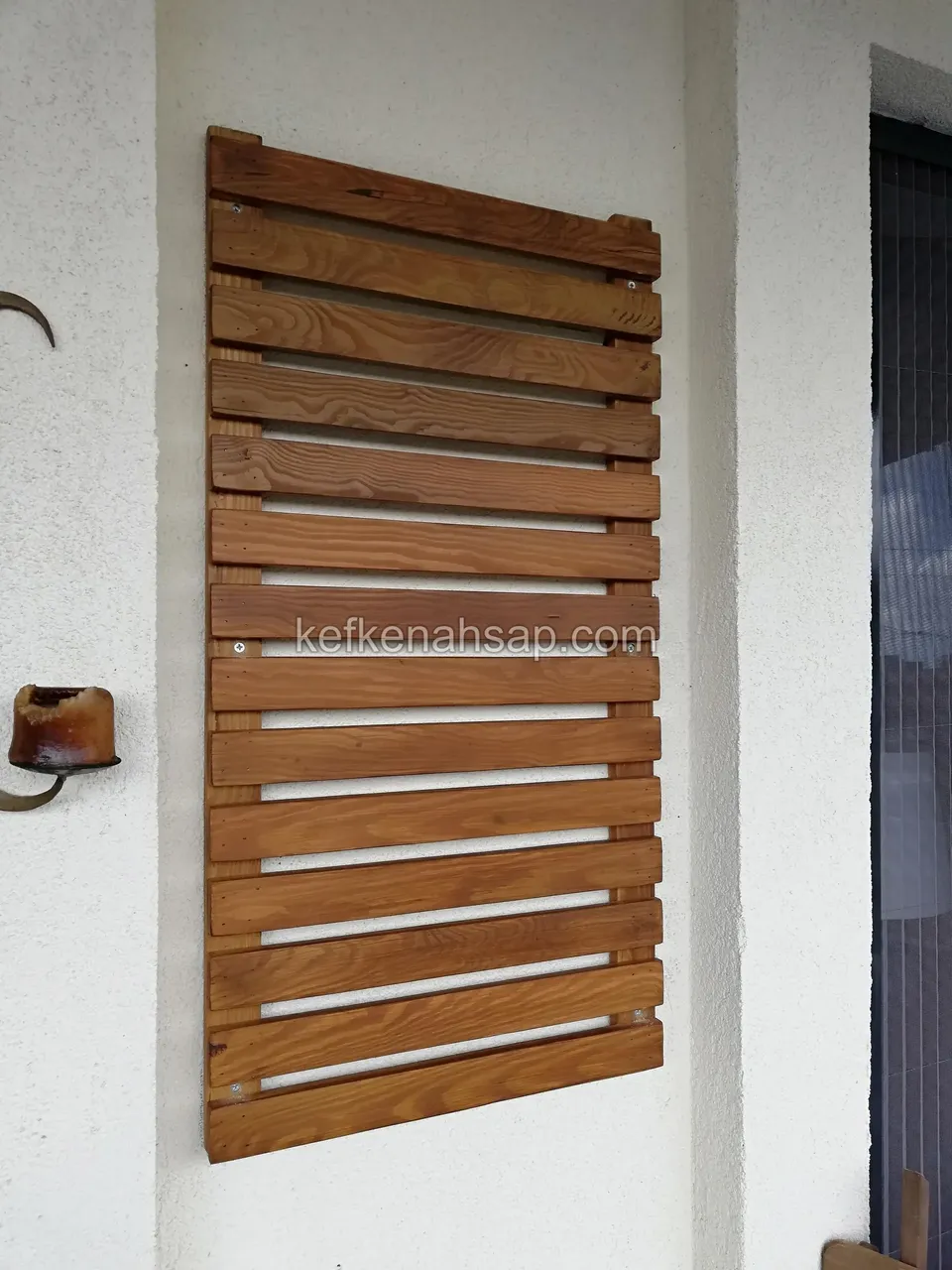 Beyaz sıvalı balkon duvarına asılmış, aralıklı yatay çıtalardan oluşan dikdörtgen açık kahve ahşap panel