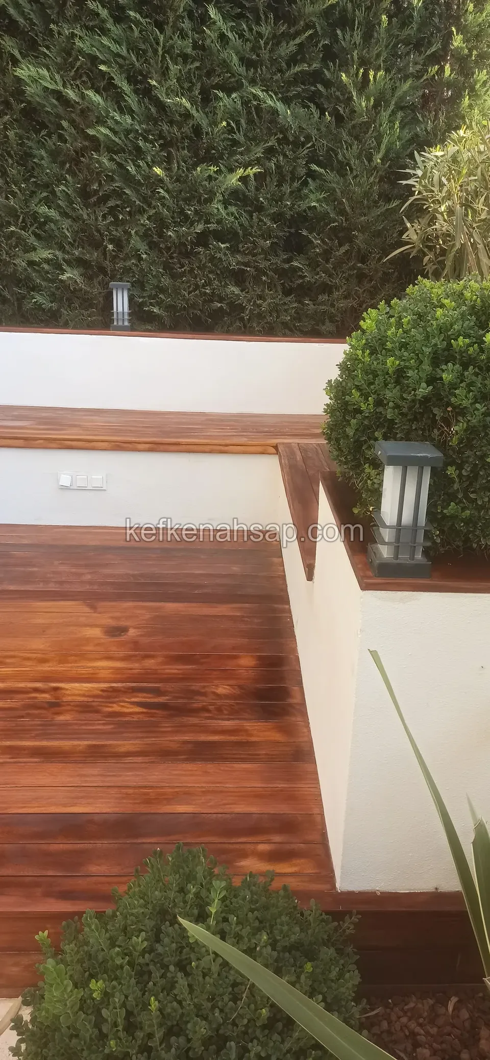 Beyaz sıvalı duvarlar arasında koyu tonlu ahşap deck ve alçak duvar üstüne oturan ahşap bank