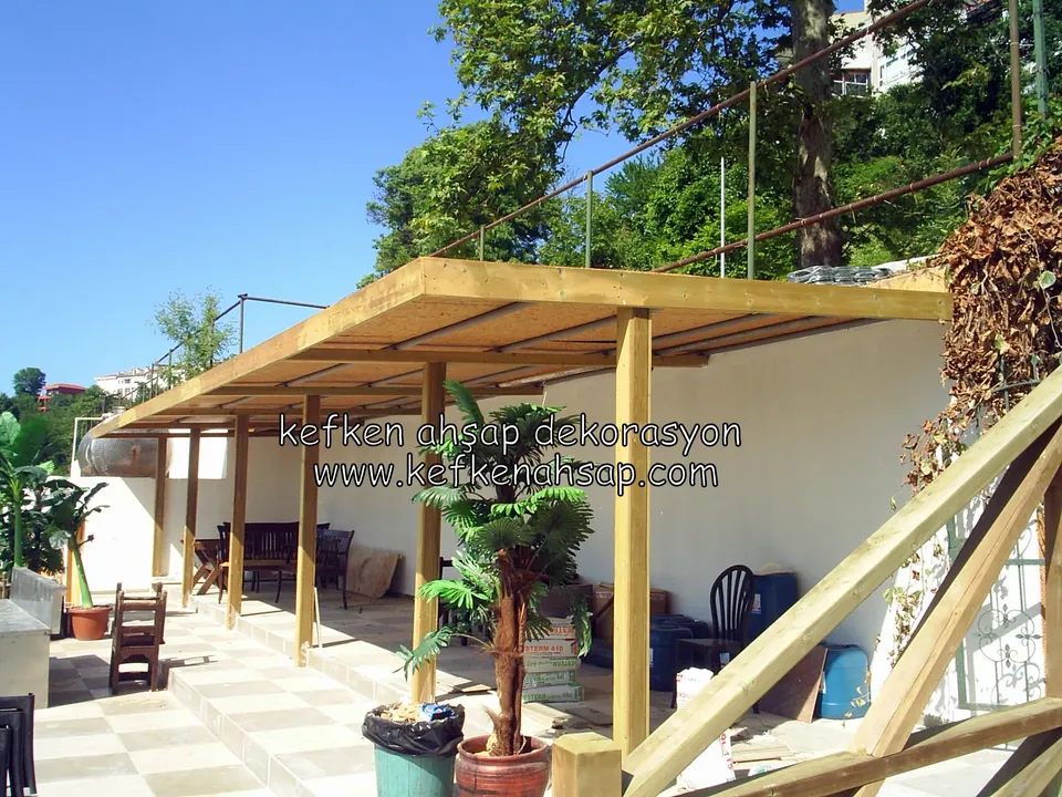 Beyaz duvarlı teras kafede açık renk ahşap pergola, altında masalar, sandalyeler ve süs bitkileri, arkada ağaçlık
