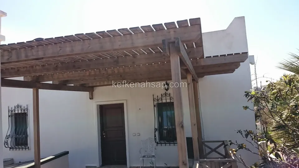 Beyaz sıvalı evin kapı önünde geniş, vernikli ahşap pergola; pencerede demir parmaklık ve yan tarafta ağaç