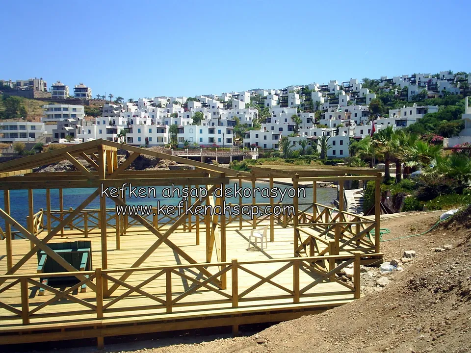 Beyaz evlerin karşısındaki deck platformunda çapraz korkuluk hattı ve üstteki çatılı kamelya, mavi deniz