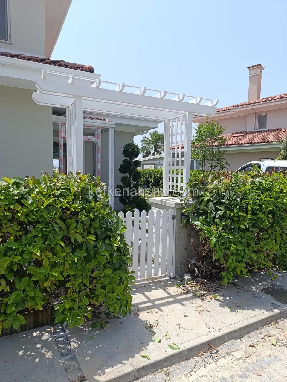 Evin girişinde beyaz boyalı pergola, kafes yan panel ve beyaz çıtalı bahçe kapısı, iki yanda çalı