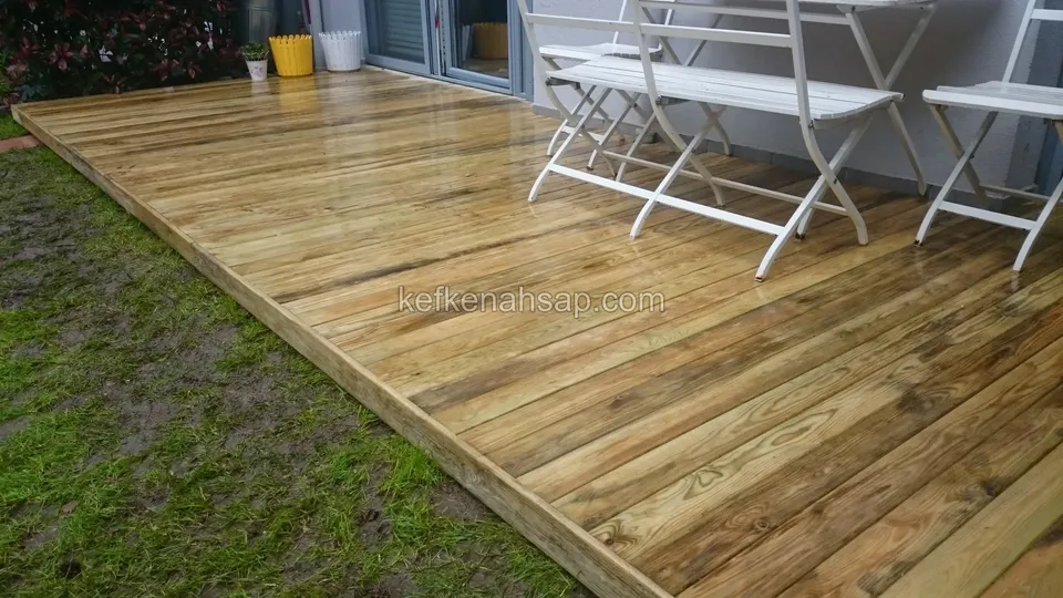 Çimenin kenarında duran, üzerinde beyaz katlanır masa ve sandalyeler bulunan açık renkli deck