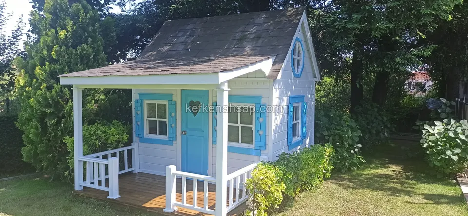 Beyaz korkuluklu verandası, mavi kapısı ve pencere kepenkleri olan oyun evi; yeşil bir bahçede
